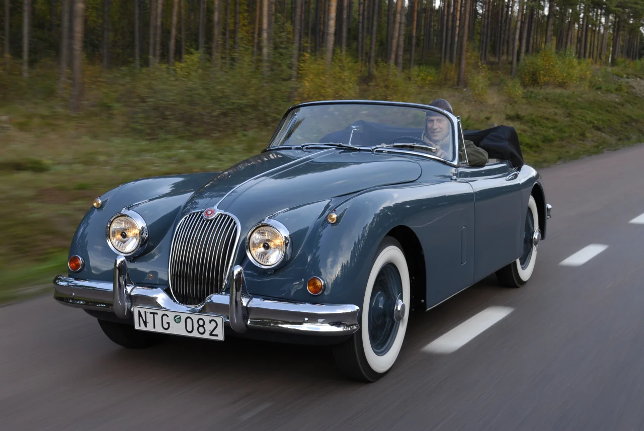 XK150 med nedfälld sufflett vid en skog, snett framifrån
